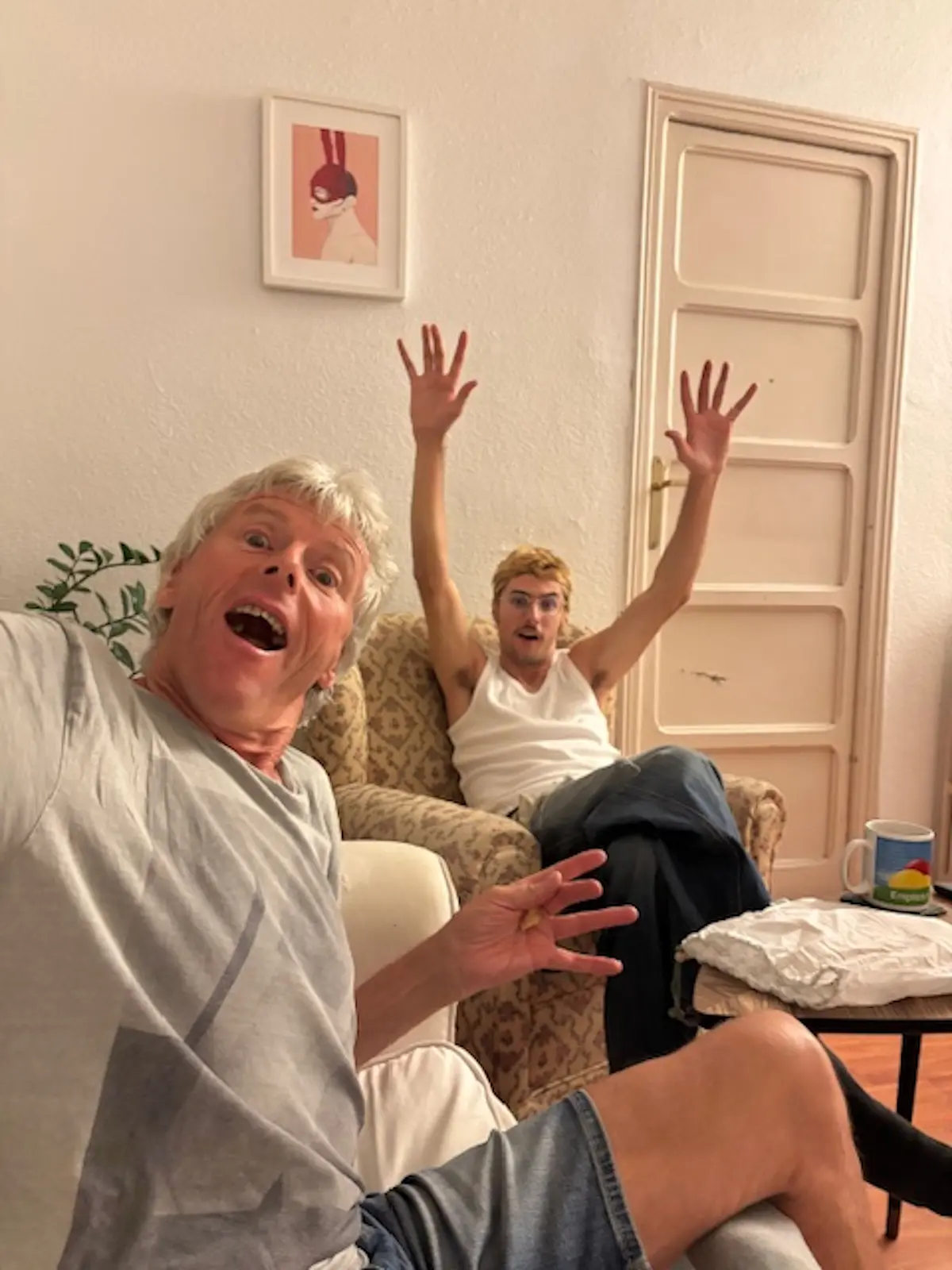 Due persone sorridenti in un soggiorno. Un uomo seduto in primo piano fa un'espressione divertente, mentre un giovane dietro di lui solleva le braccia con entusiasmo. Nel fondo si vede un quadro appeso al muro.