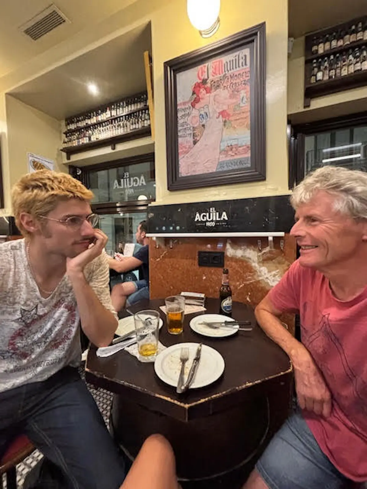 Due uomini seduti a un tavolo in un bar, uno con capelli biondi corti e occhiali, l'altro con capelli bianchi ricci. Entrambi sorridono mentre conversano, con piatti e bicchieri di birra sul tavolo. Sullo