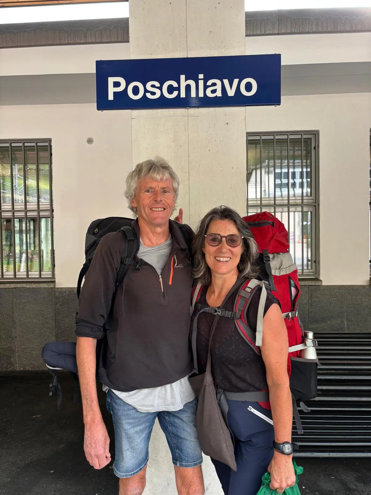Due escursionisti sorridenti in stazione a Poschiavo, con zaini e attrezzatura da trekking.