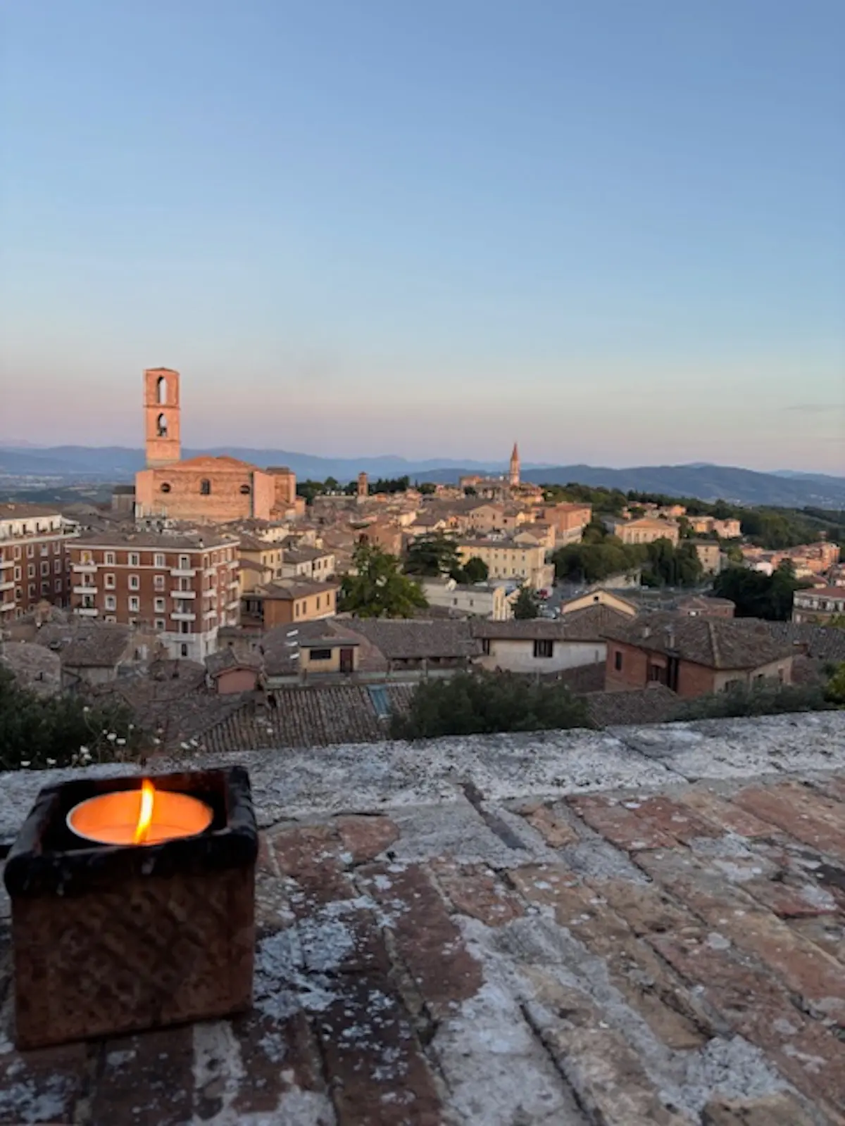 Vista panoramica di una città con edifici storici, un campanile e colline sullo sfondo. In primo piano, una candela accesa su una superficie di mattoni. Il cielo mostra tonalità di blu e rosa al tramonto.