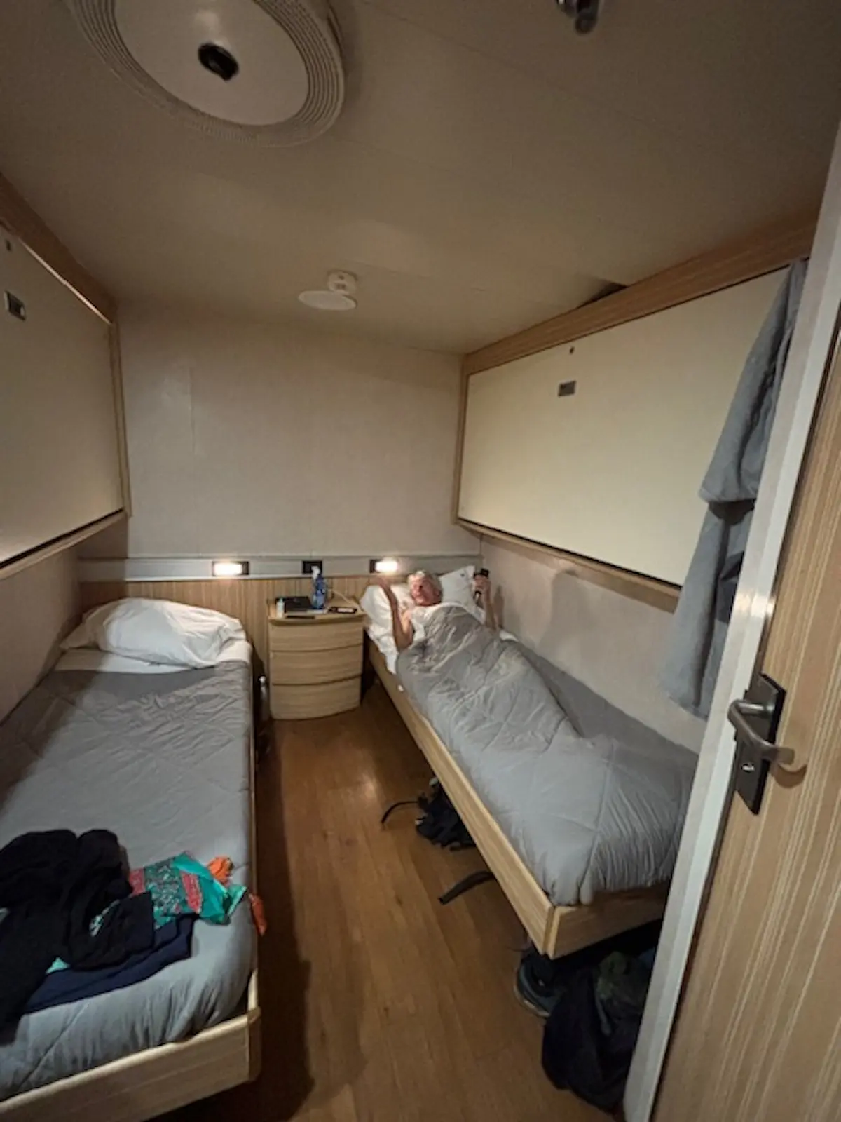 Stanza di una cabina con due letti singoli. Una persona è sdraiata su un letto, mentre l'altro letto è vuoto. Le pareti sono chiare e ci sono armadietti sopra i letti. Una porta aperta mostra l'ingresso alla cab