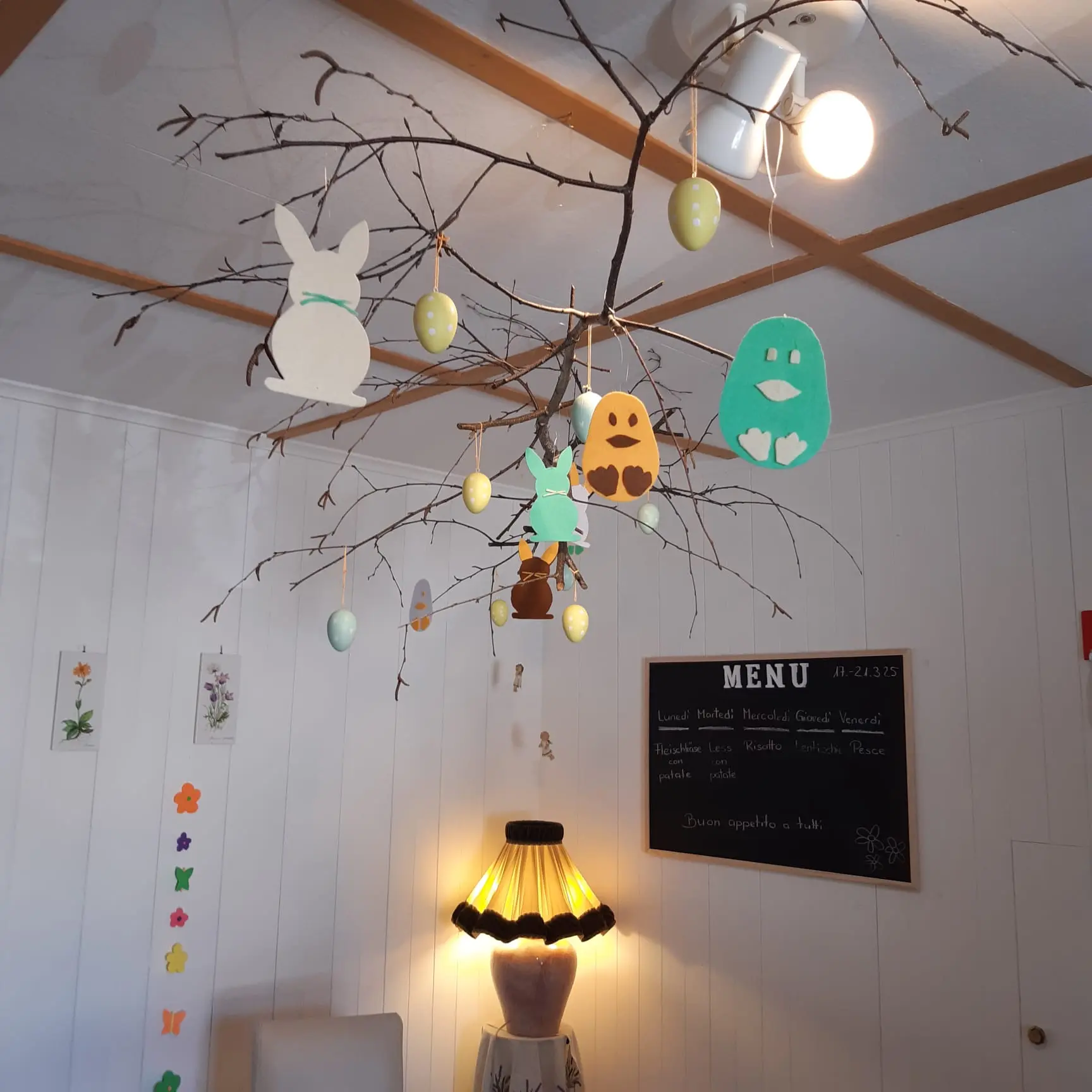 Rami Decorati con Decorazioni di Pasqua, Inclusi Coniglietti e Uova Colorate Sospesi, Sotto una Lampada e una Lavagna con un Menu.