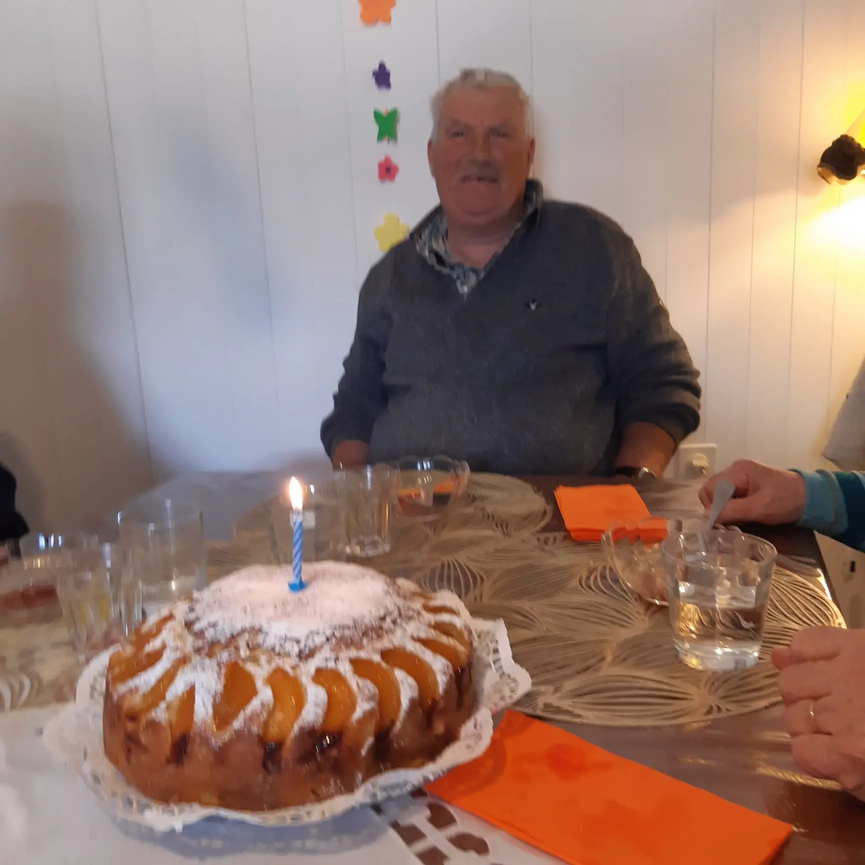 Uomo sorridente seduto a un tavolo con un dolce decorato con una candela accesa. Il tavolo è apparecchiato con bicchieri e tovaglioli colorati. Sullo sfondo si vedono decorazioni floreali.