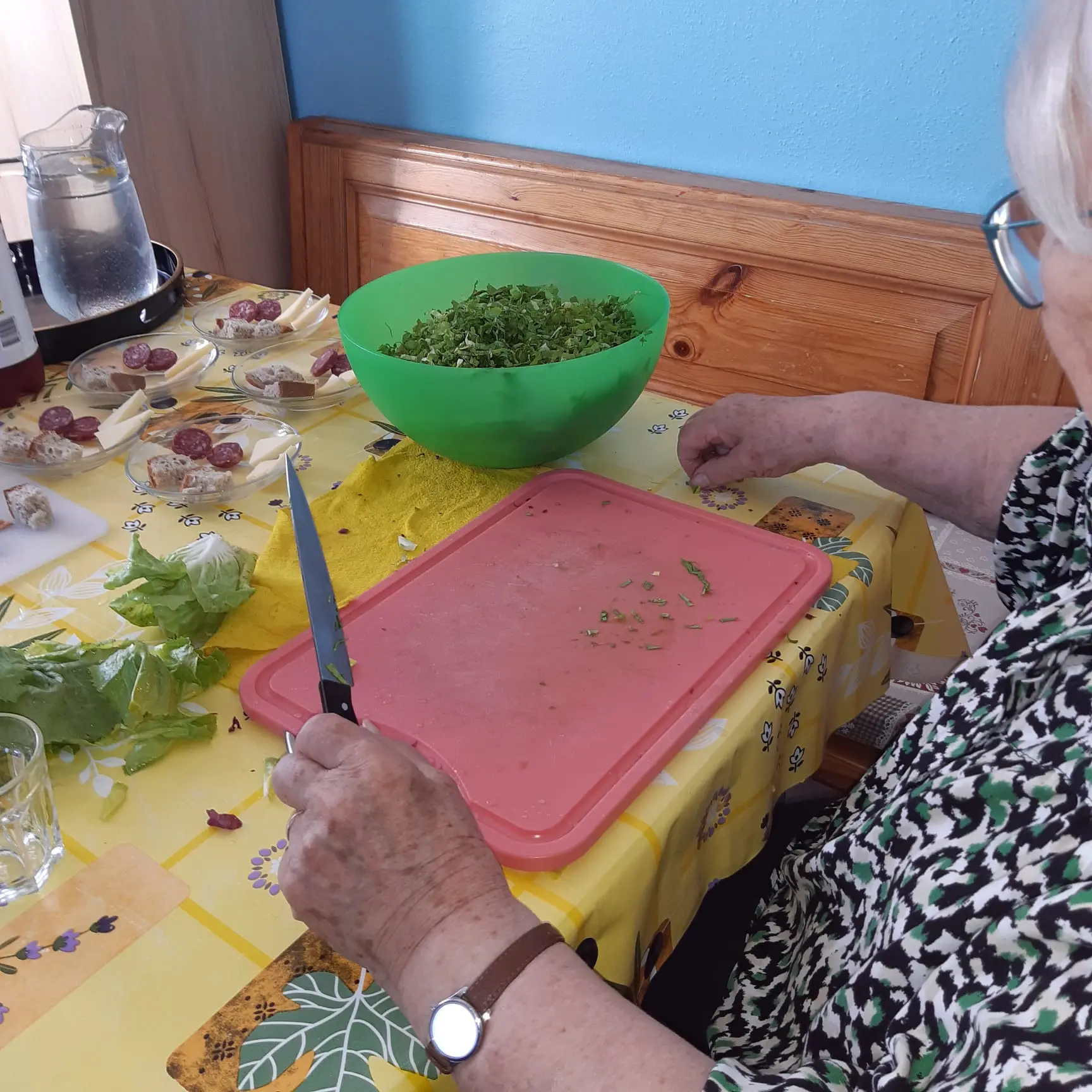 Una persona anziana sta tagliando verdure su un tagliere rosso mentre prepara un'insalata in una ciotola verde. Sul tavolo ci sono altri ingredienti e un bicchiere.