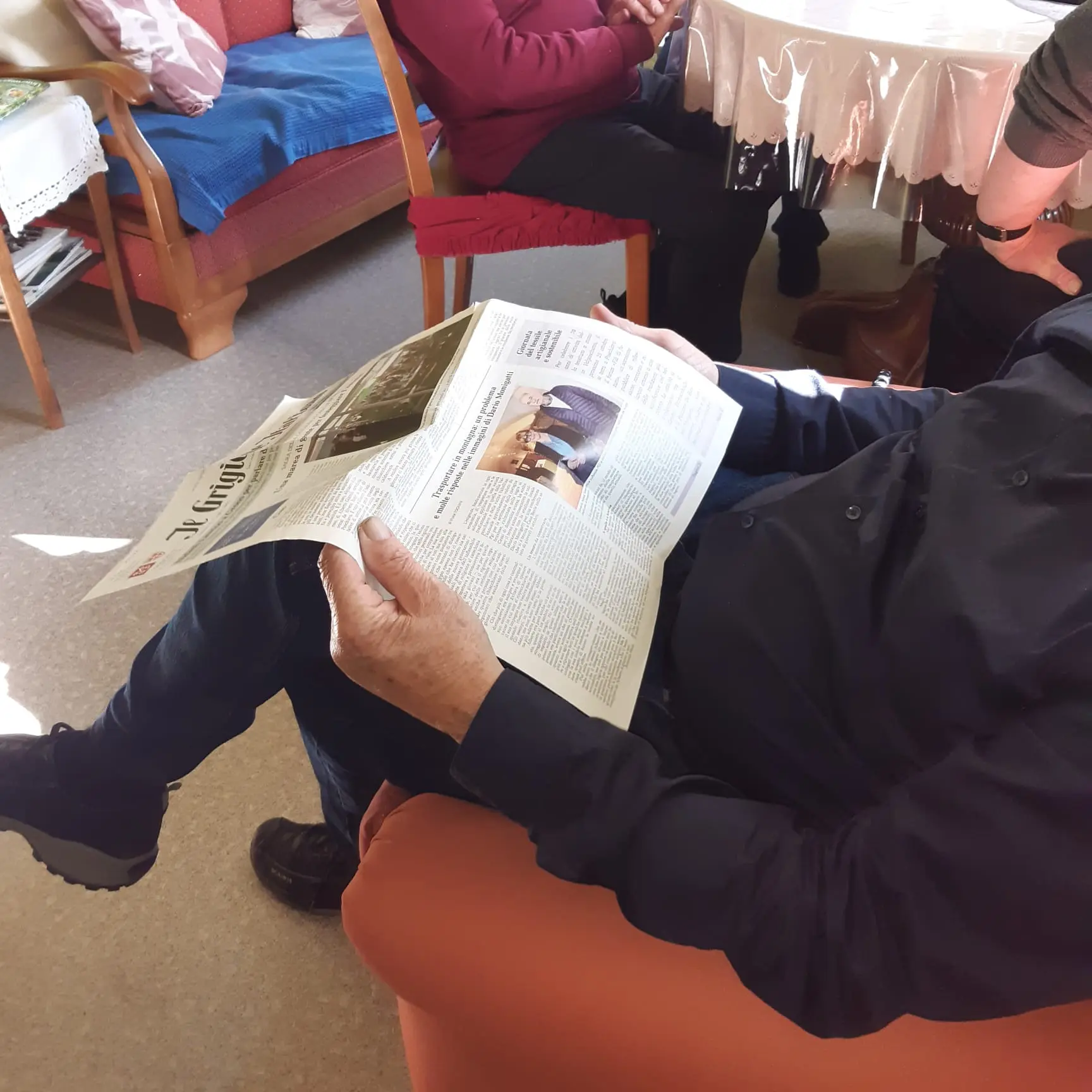 Un uomo seduto su un divano legge un giornale, con persone sedute in background. La luce illumina la sua mano e il giornale.