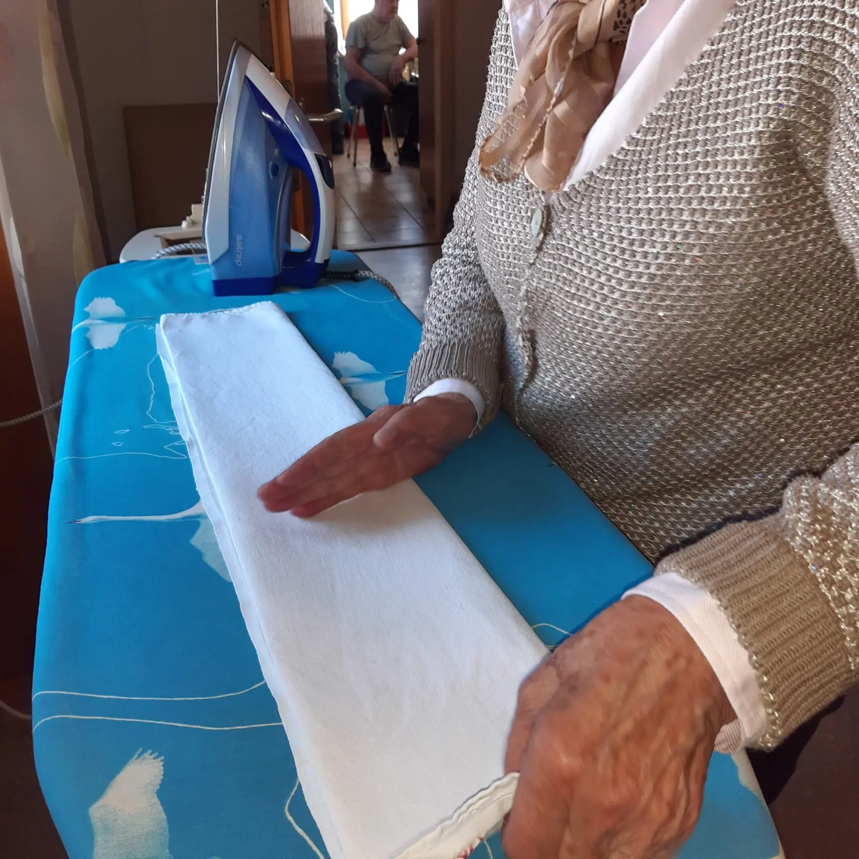 Una persona anziana sta stirando una camicia bianca su un asse da stiro, mentre un ferro da stiro blu è posizionato accanto. Il tessuto dell'asse presenta un motivo azzurro.