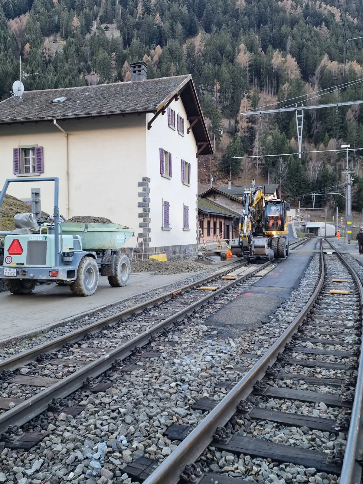 Treno in fase di costruzione lungo i binari, con un escavatore e un rimorchio carico di materiali. Sullo sfondo, una stazione ferroviaria e un edificio