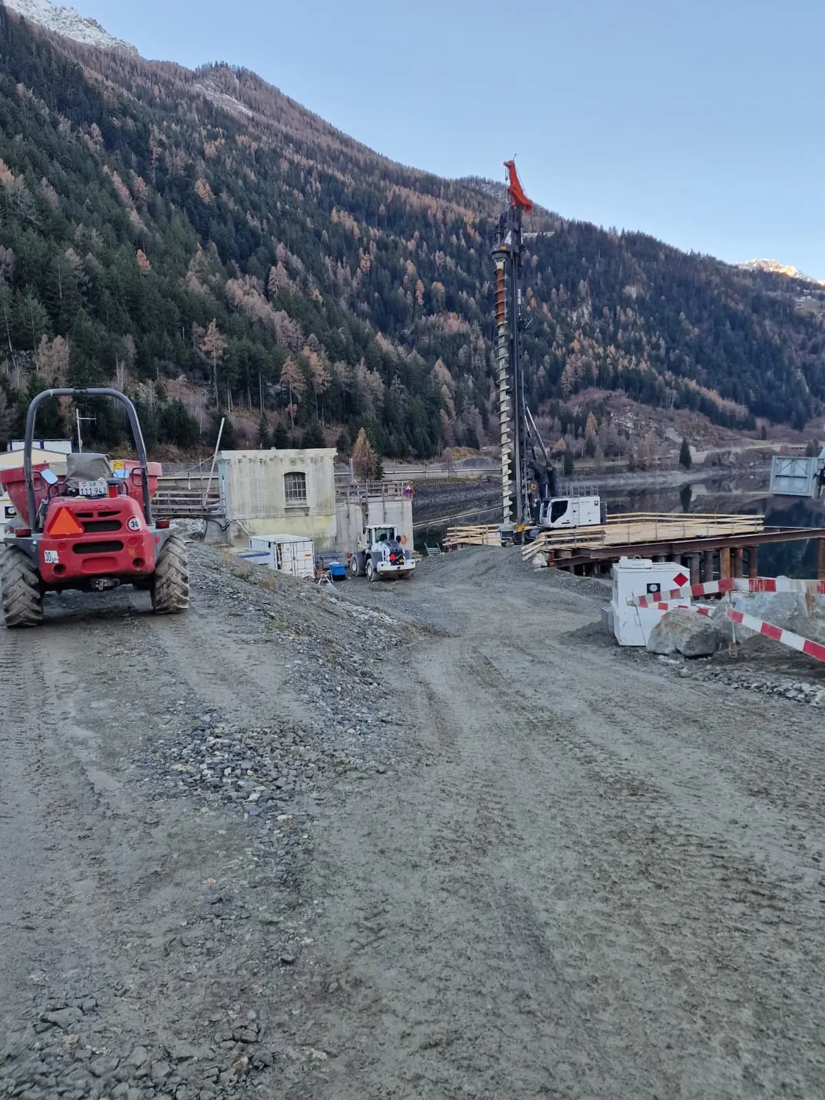 Un cantiere di costruzione montano con macchinari pesanti, inclusi un trattore rosso e una torre di perforazione. Ci sono strutture e materiali da costruzione lungo