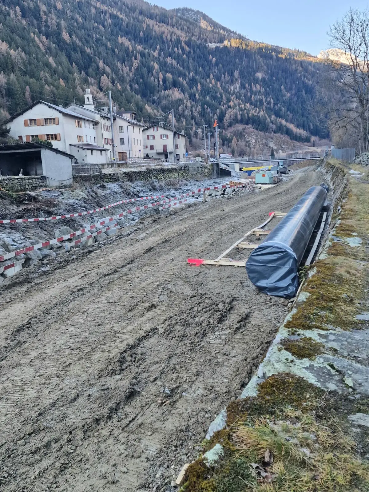 Cantiere stradale in una vallata montana, con terreno smosso e una tubazione nera avvolta. Sullo sfondo, edifici di montagna e alber