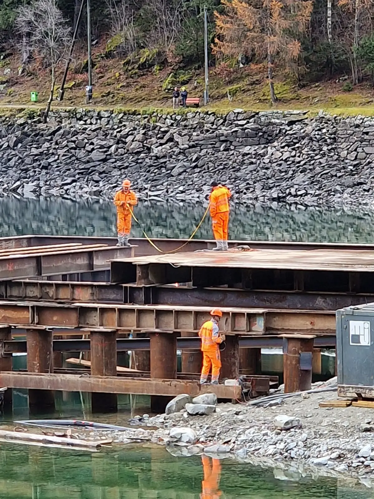 Operai vestiti in tute arancioni lavorano su una piattaforma metallica vicino a un corpo d'acqua. Sullo sfondo si vedono alberi e una scogliera