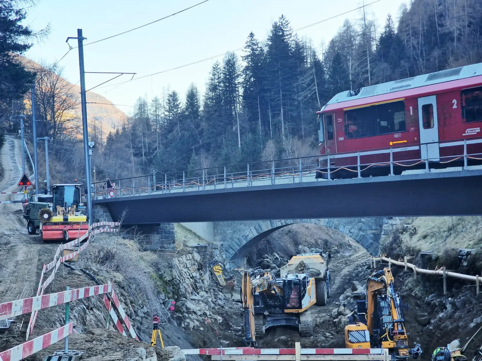 Treno rosso attraversa un ponte mentre scavatrici e attrezzature da cantiere lavorano sotto di esso, in un'area in fase di costruzione circondata