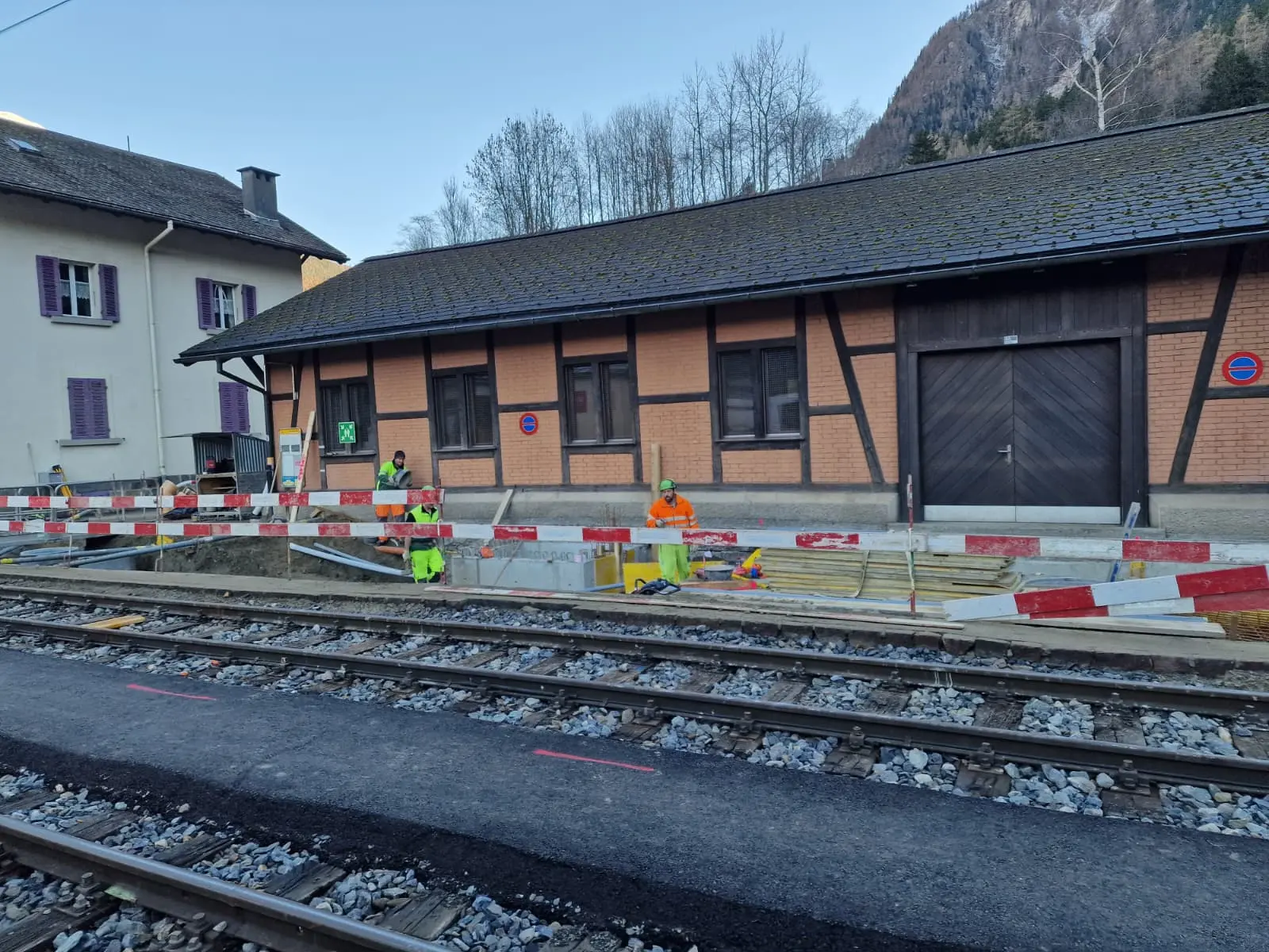 Operai stanno effettuando lavori di riparazione vicino ai binari di una stazione ferroviaria. In primo piano si vedono segnaletica di sicurezza e ostacoli, mentre sul