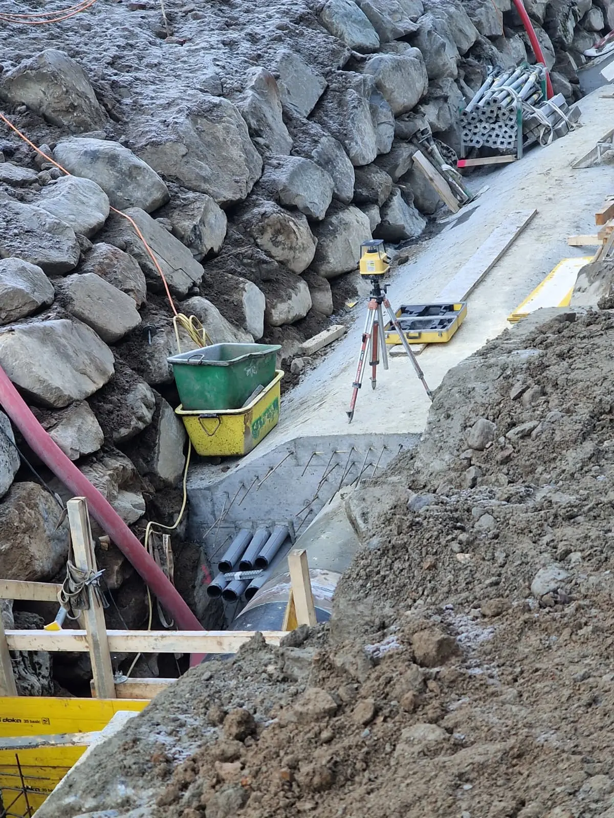 Un'area di costruzione con un letto di ghiaia e una fila di rocce. Sono visibili attrezzature per la misurazione, come un livello topografico, e