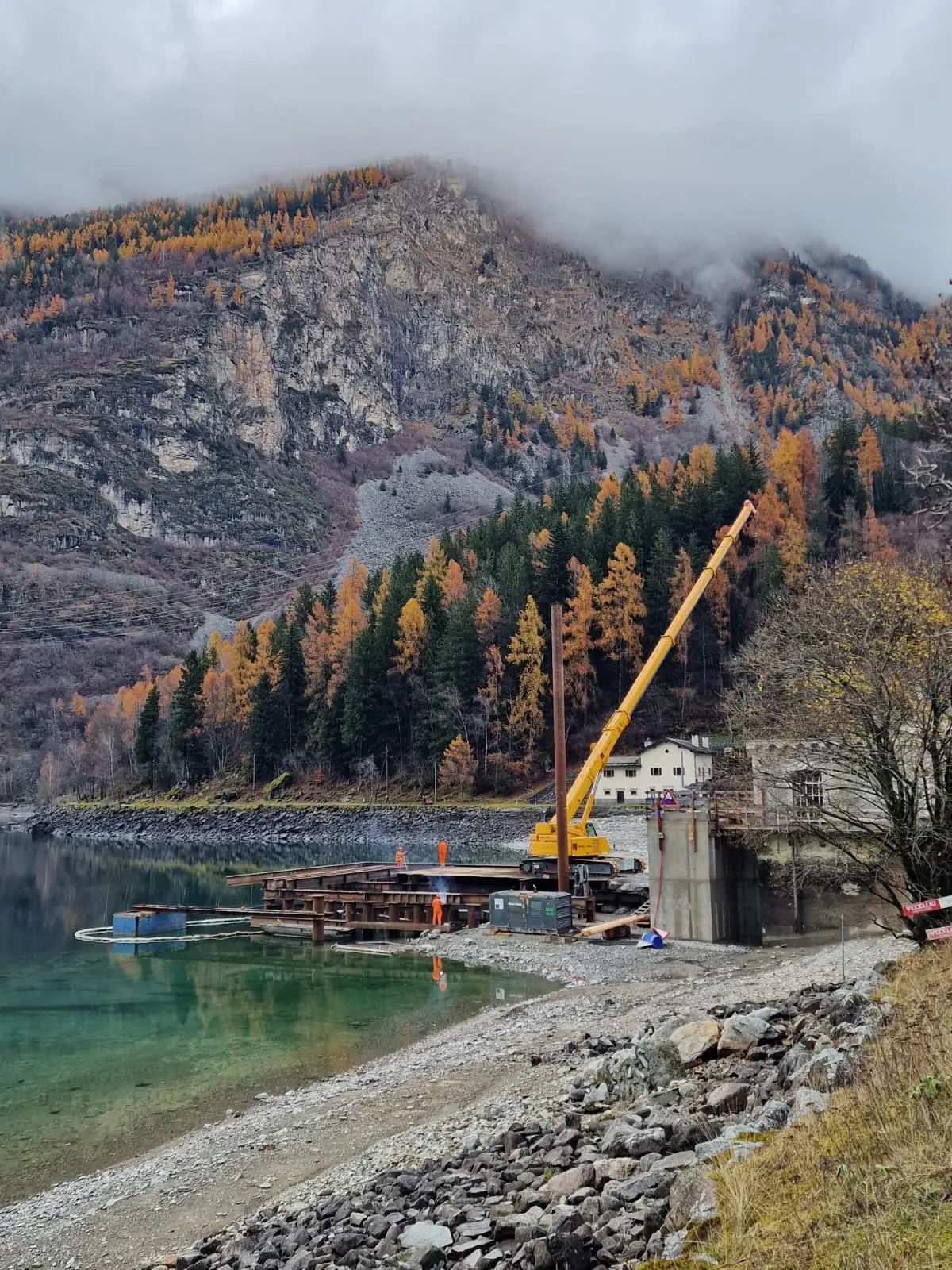 Un cantiere sul bordo di un lago, con una gru gialla che si erge su una piattaforma di legno. Sullo sfondo, montagne con alberi autunnali e