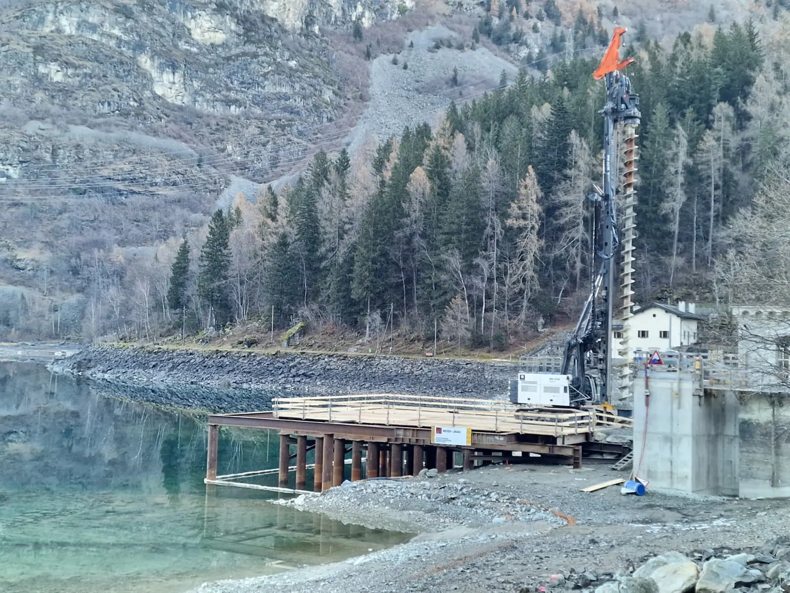 Un cantiere di costruzione vicino a un lago, con una torre di perforazione e una passerella in legno. Sullo sfondo, alberi di conifere e montagne.