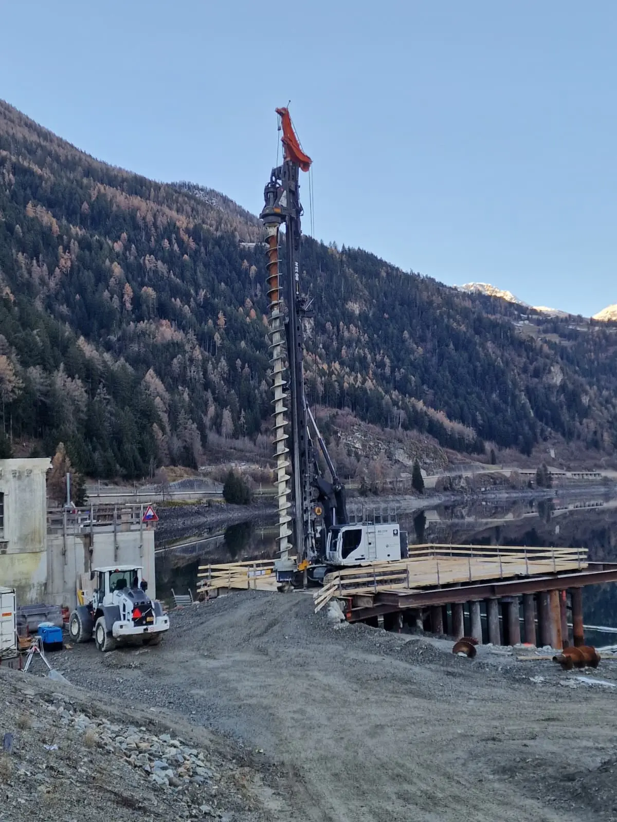 Una grande macchina perforatrice si erge sopra un cantiere, circondata da montagne e alberi con foglie autunnali. Il cantiere ha una rampa di accesso