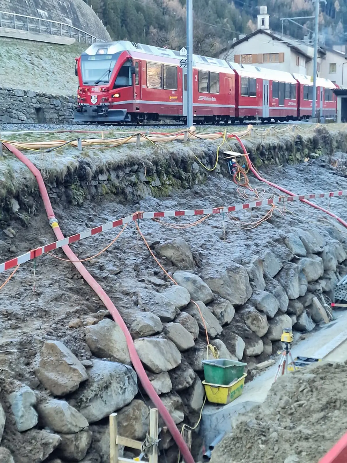 Un treno rosso passa lungo un tratto di binario, con lavori di costruzione in primo piano che mostrano un muro di pietre e tubi arancioni. Sullo sf