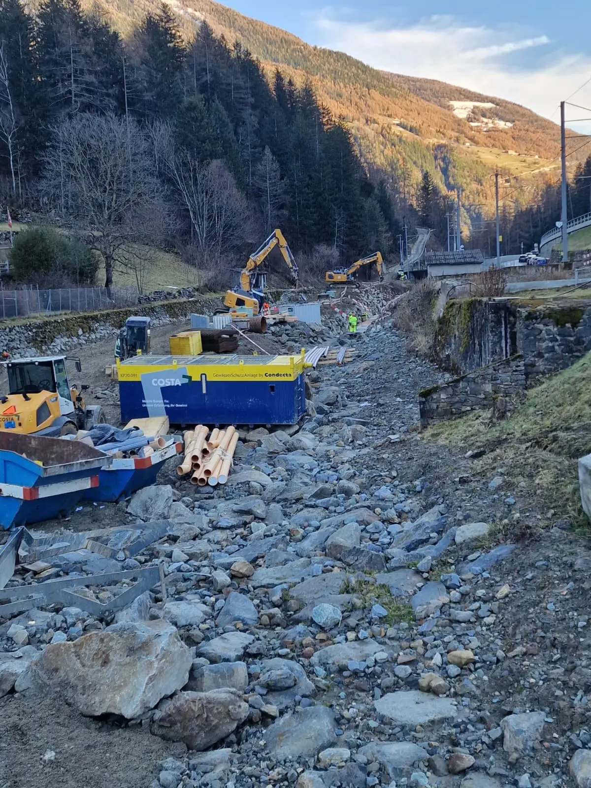 Cantiere in una zona montuosa con macchinari pesanti, camion e tubi sistemati lungo un letto di roccia. Sullo sfondo si vedono alberi e