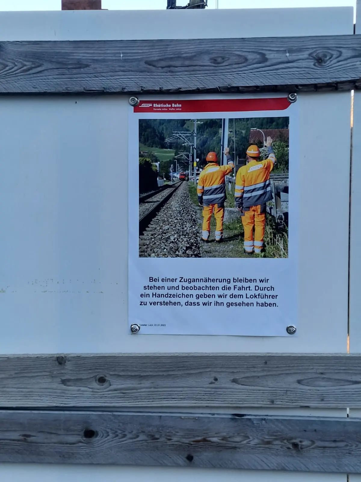 Cartello informativo che indica l'importanza di fermarsi e osservare i treni durante l'approccio ai binari, con due operai in uniforme arancione che osservano la dire