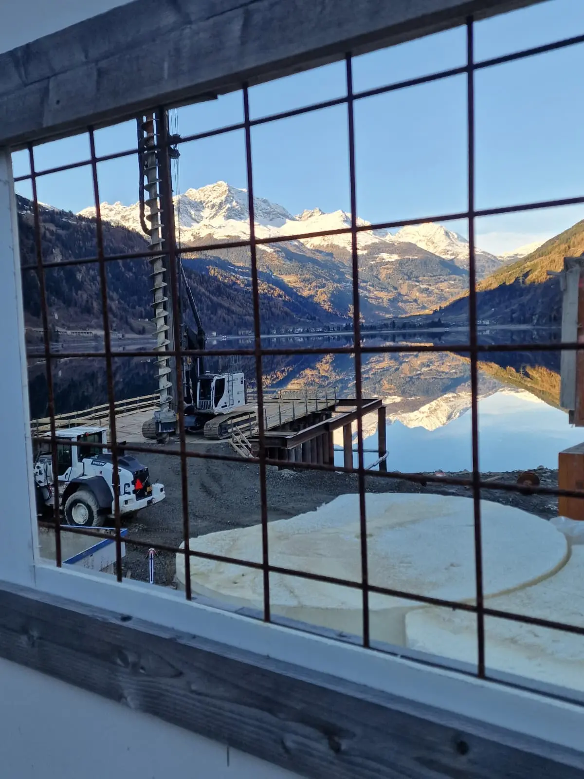 Vista attraverso una grata che mostra un lago tranquillo circondato da montagne innevate. Un ponte di legno e attrezzature da costruzione sono visibili in primo piano, mentre il