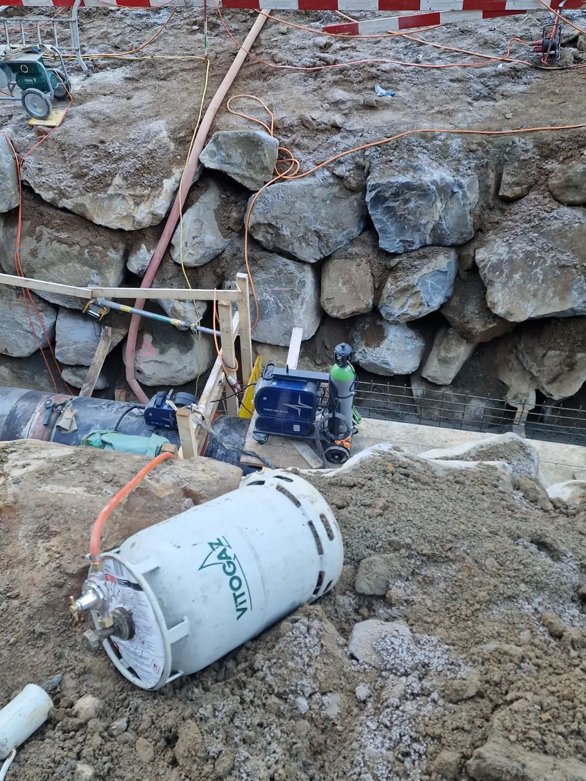 Un cantiere edile con rocce grandi sullo sfondo, attrezzature e materiali da costruzione sparsi, tra cui un serbatoio e tubazioni.