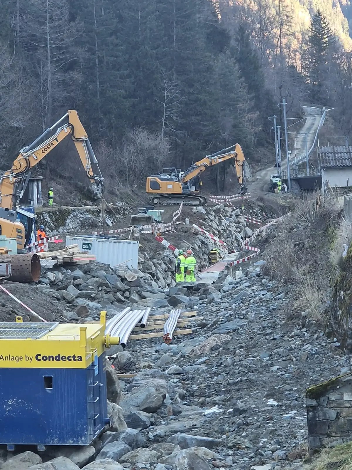 Un cantiere stradale con escavatori e operai che lavorano su un terreno seminato di pietre. Si vedono tubi metallici e segnali di sicurezza lungo il