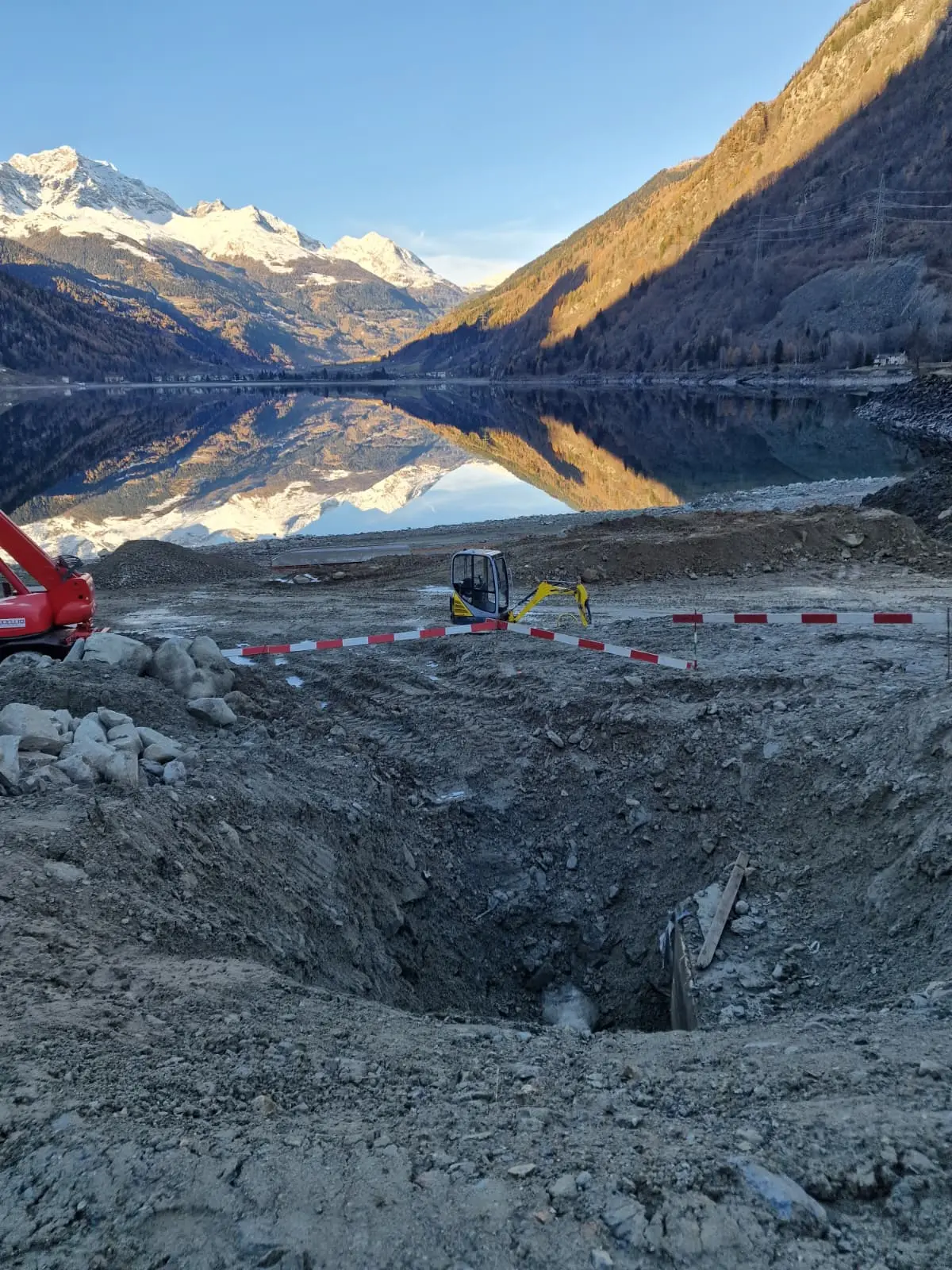 Sito di scavo con una grande buca nel terreno, circondato da transenne rosse e bianche. Sullo sfondo, montagne coperte di neve si riflet