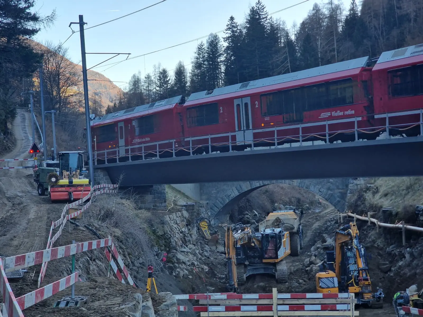Un treno rosso passa sopra un ponte mentre si svolgono lavori di costruzione e scavi sottostanti. Macchinari pesanti sono visibili sulla terra in basso, circond