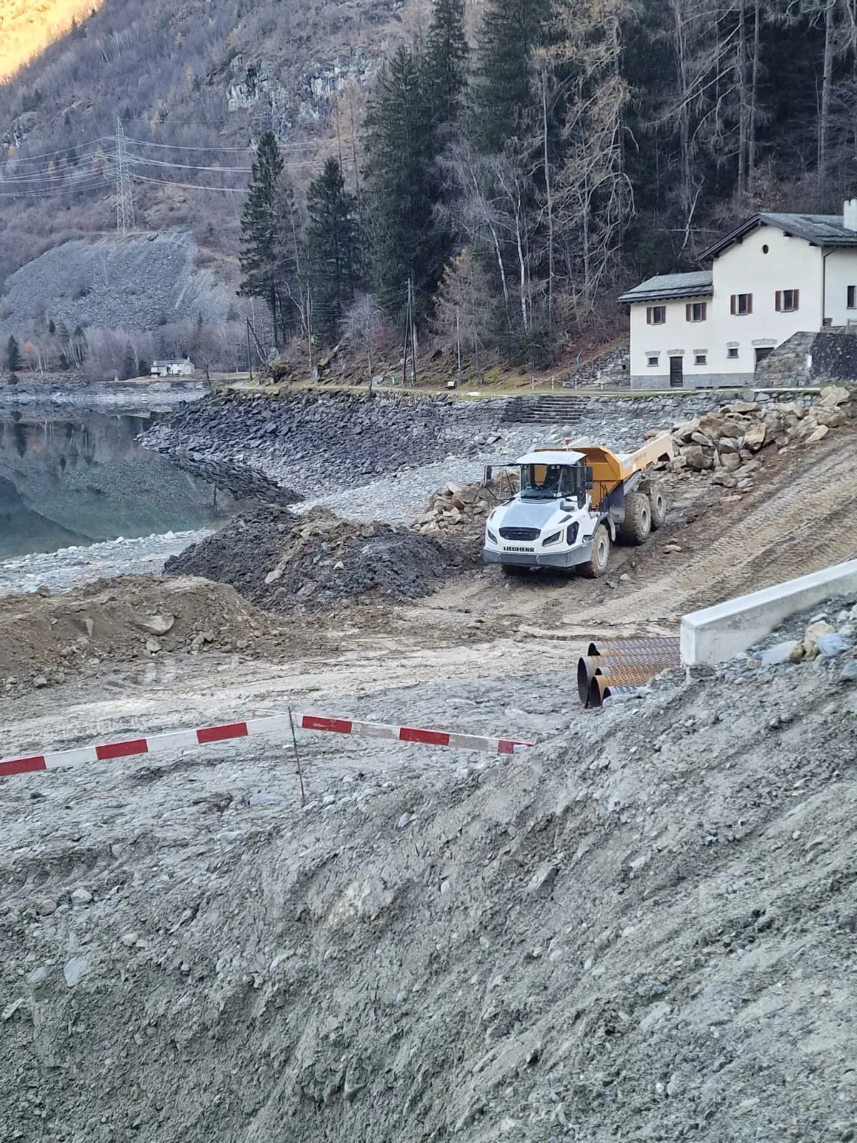 Un dumper giallo lavora su un terreno di cantiere vicino a un lago, con alberi sullo sfondo e una piccola costruzione bianca. La superficie è ster
