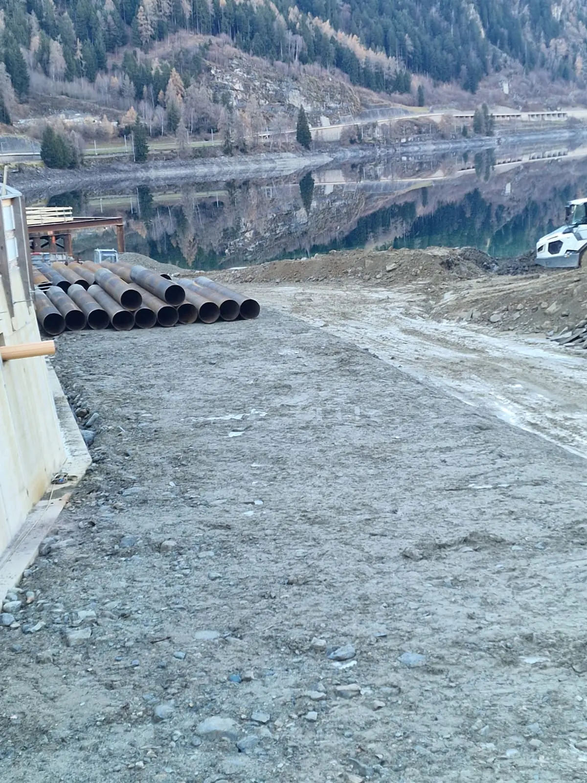 Un cantiere in una zona montuosa con tubi di metallo sistemati su un terreno ghiaioso vicino a un lago. Le montagne si riflettono nell'acqua.
