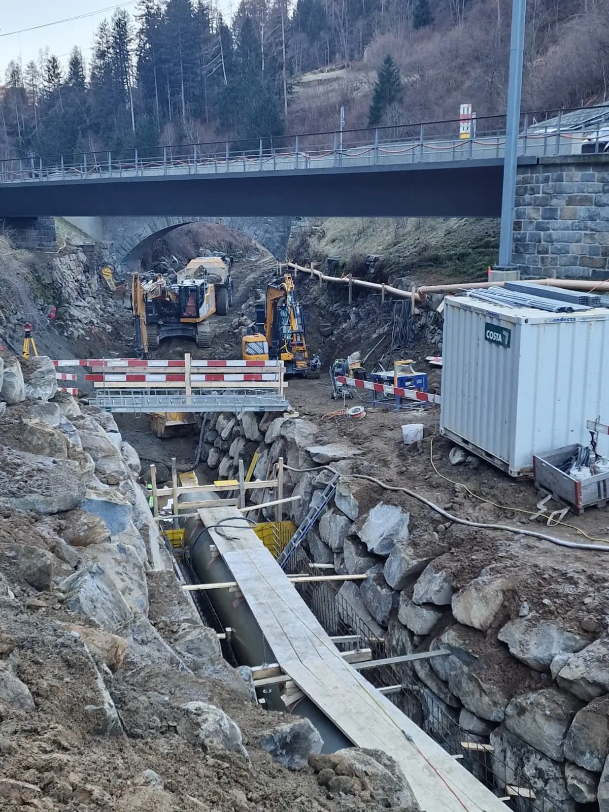 Cantiere in fase di scavo con macchinari pesanti, tubazioni e materiali da costruzione, accompagnato da un ponte sovrastante.