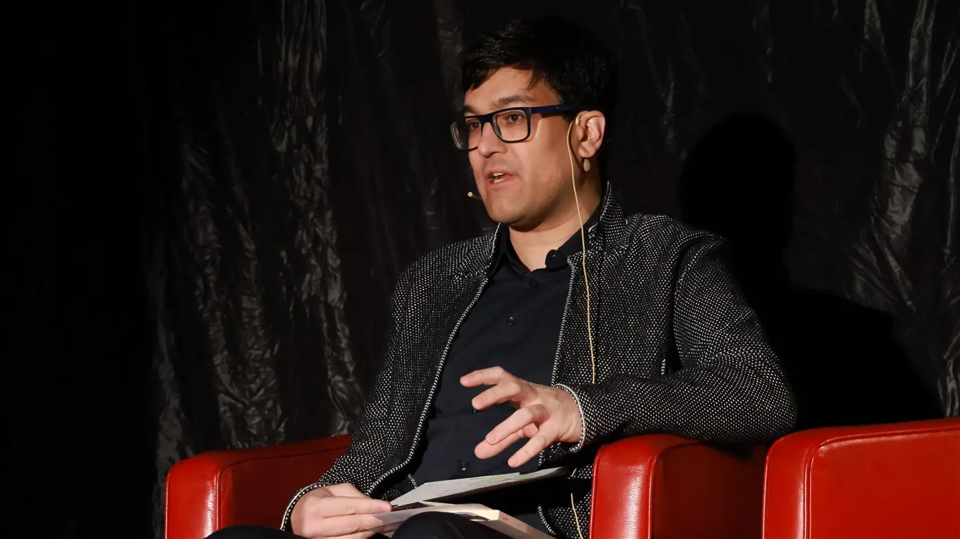 Un uomo con occhiali e una giacca elegante parla in un microfono, seduto su una sedia rossa. Sullo sfondo, un tessuto nero.