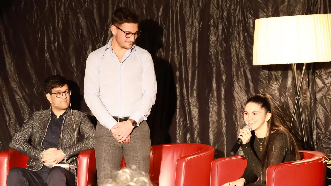 Tre persone sedute su poltrone rosse in un evento. Due uomini osservano una donna che parla, mentre una lampada illuminata si trova dietro di loro. Il fondo è di