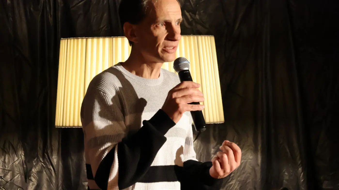 Un uomo con un maglione a righe sostiene un microfono mentre parla, con una lampada sullo sfondo che illumina la scena.