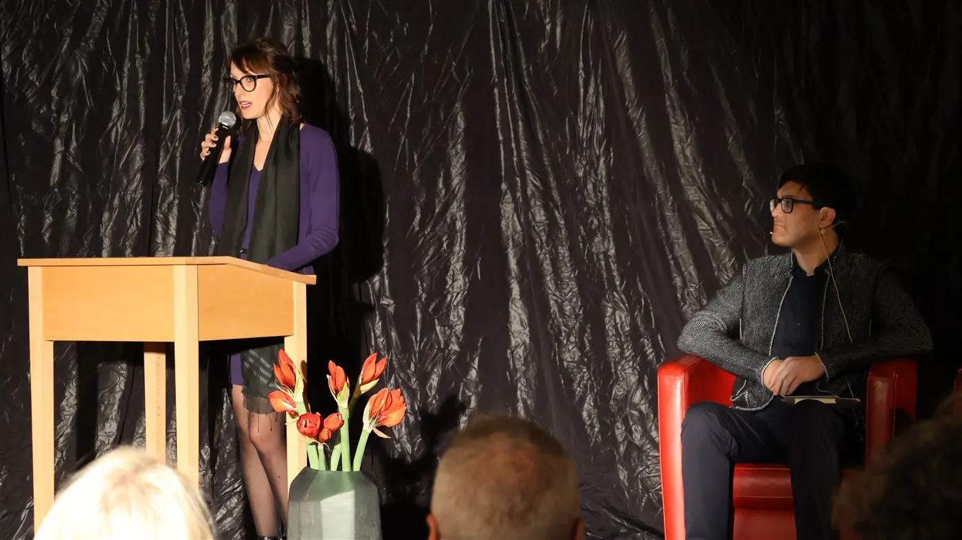 Una donna in piedi al microfono, con occhiali e abbigliamento scuro, parla a un pubblico. Seduto accanto a lei su una poltrona rossa, un