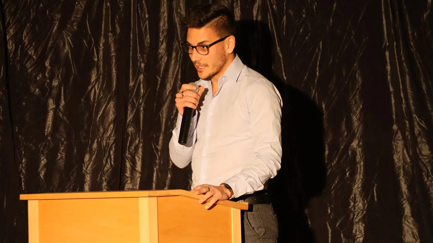 Un uomo con occhiali e camicia bianca parla al microfono, appoggiato a un leggio su uno sfondo scuro.