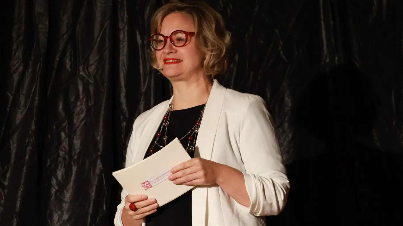 Una donna con occhiali rossi e capelli biondi ricci indossa una giacca bianca e tiene un documento in mano. Sta parlando di fronte a uno sfondo scuro