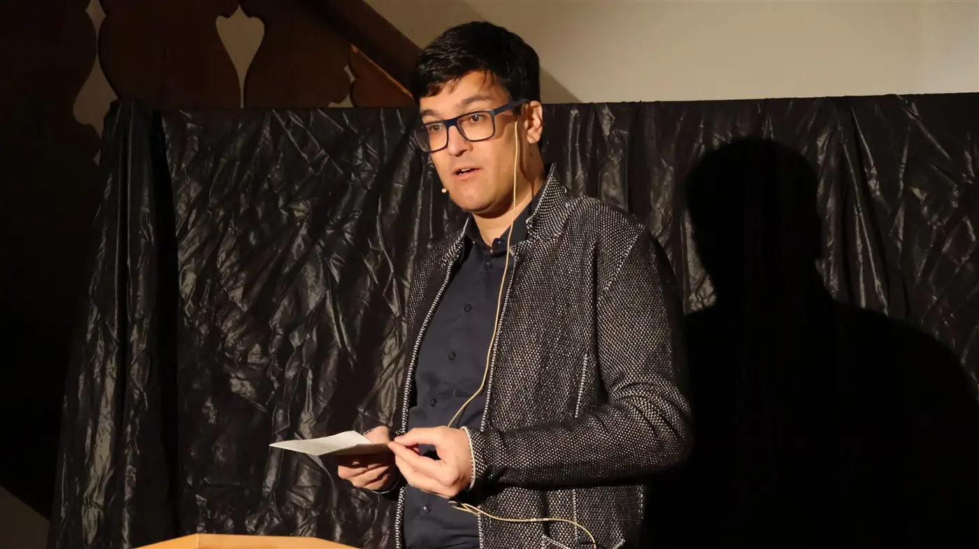 Un uomo in giacca scura parla davanti a un pubblico, con una nota in mano. Sullo sfondo, un drappo nero crea un'atmosfera di concentrazione.