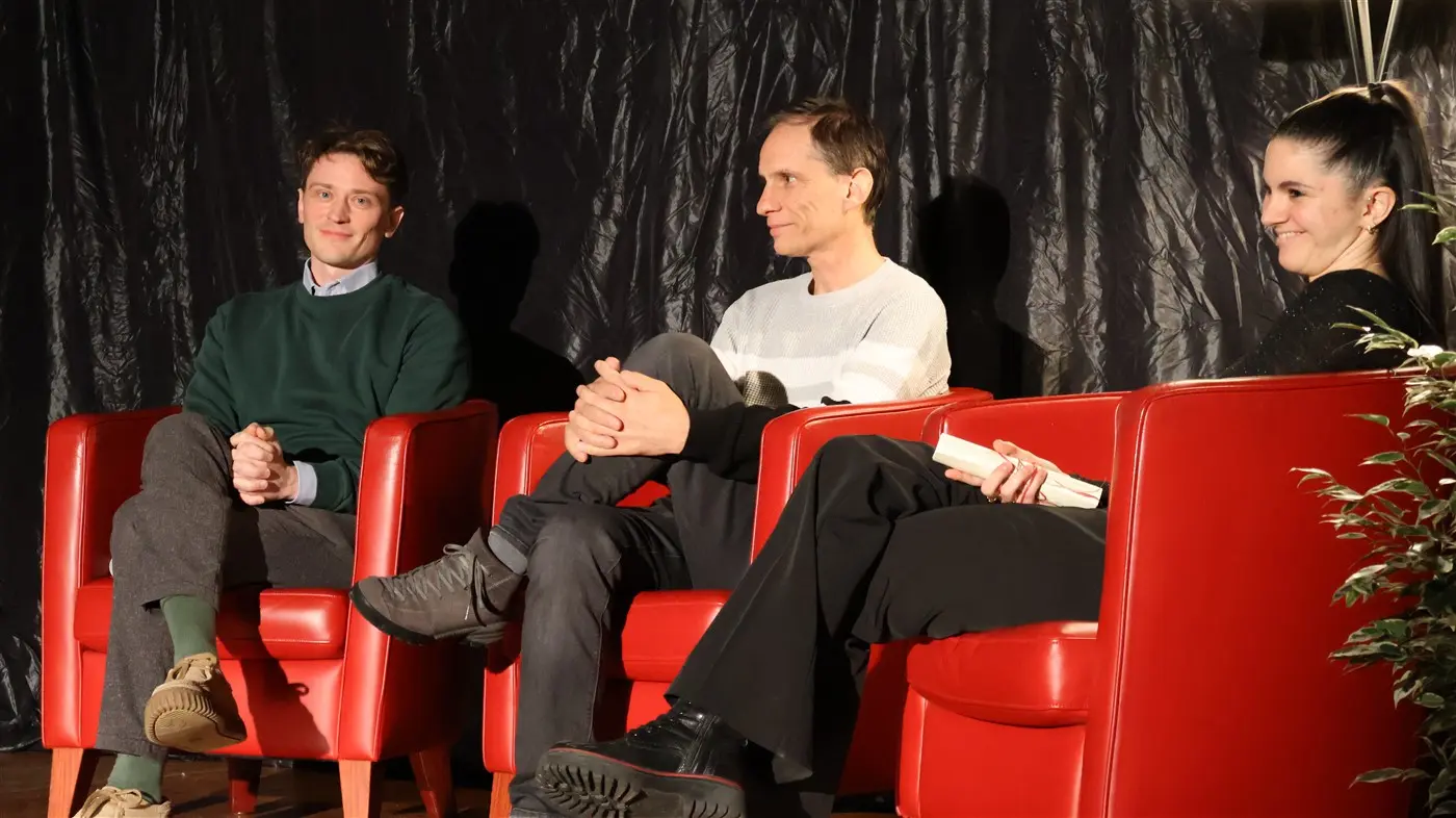 Tre persone sedute su poltrone rosse durante un'intervista. Due uomini e una donna sorridono mentre si scambiano idee. Sullo sfondo, una tenda nera