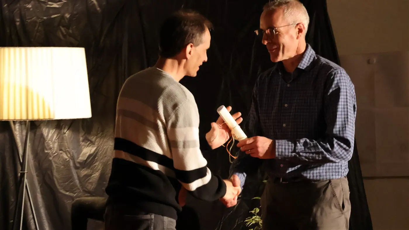 Un uomo con una maglietta a righe stringe la mano a un altro uomo in camicia, mentre consegna un rotolo di carta. Sullo sfondo si vede una lampada