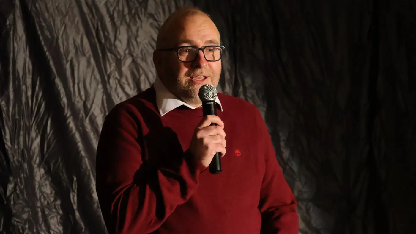 Uomo con occhiali e maglione rosso parla al microfono, illuminato da una luce soft su uno sfondo scuro.