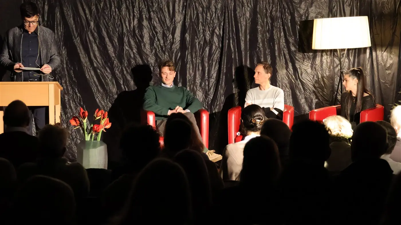 Un evento culturale con quattro relatori, tre seduti su poltrone rosse e uno in piedi al leggio. Sullo sfondo, una parete nera e una lampada