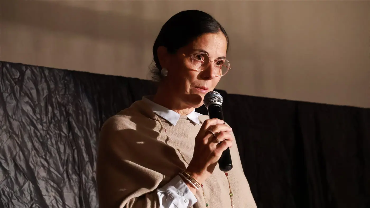 Una donna con occhiali e orecchini large parla al microfono, indossando un poncho chiaro su una camicia bianca. Sullo sfondo c'è un drapp