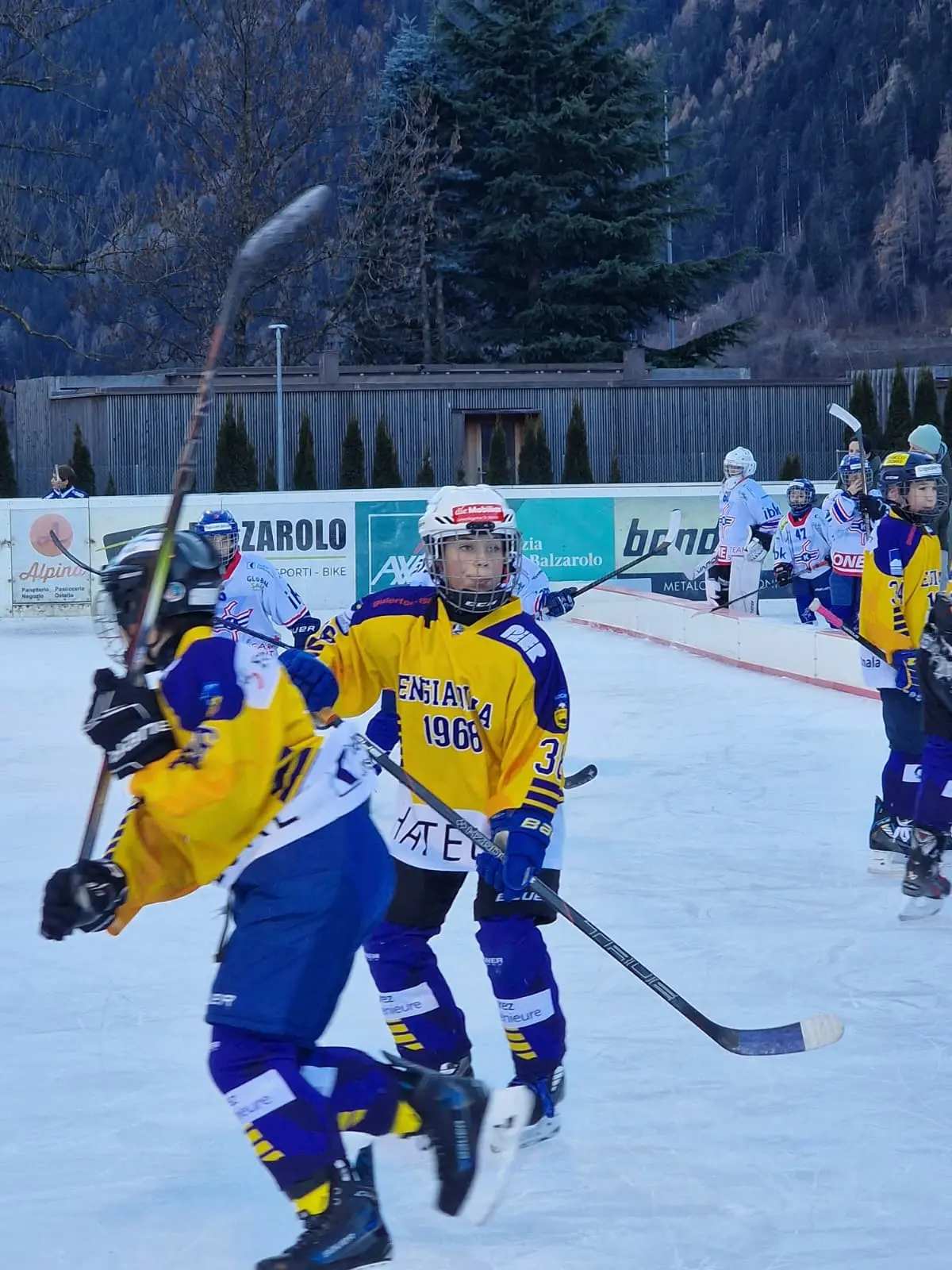 Giocatori di hockey su ghiaccio in azione su un campo all'aperto, indossando divise gialle e blu. Si nota un giocatore con il numero 31 che