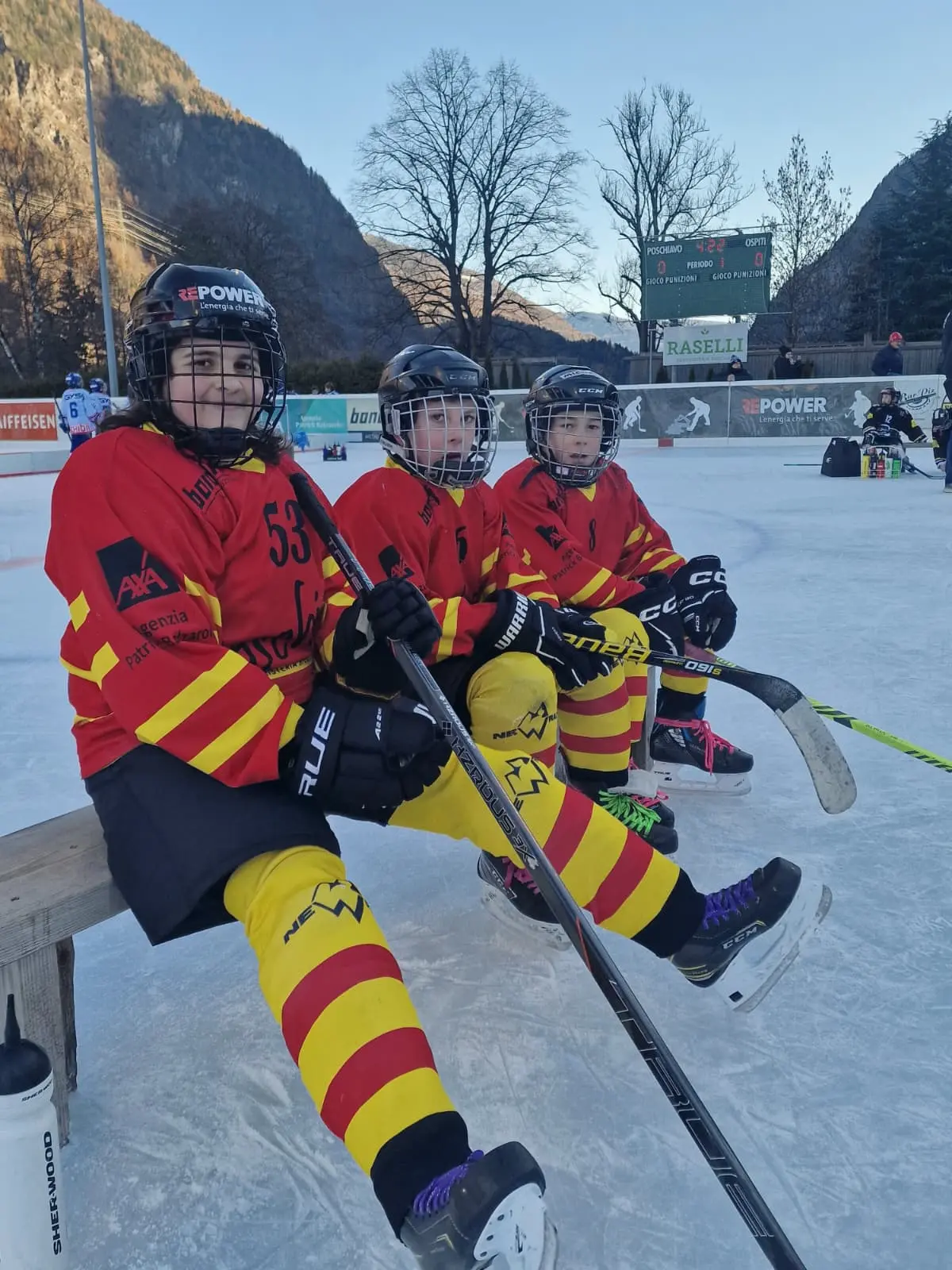 Tre giovani giocatori di hockey su ghiaccio seduti su una panchina, indossando uniformi rosse e gialle. Sorridono e tengono i bastoni da