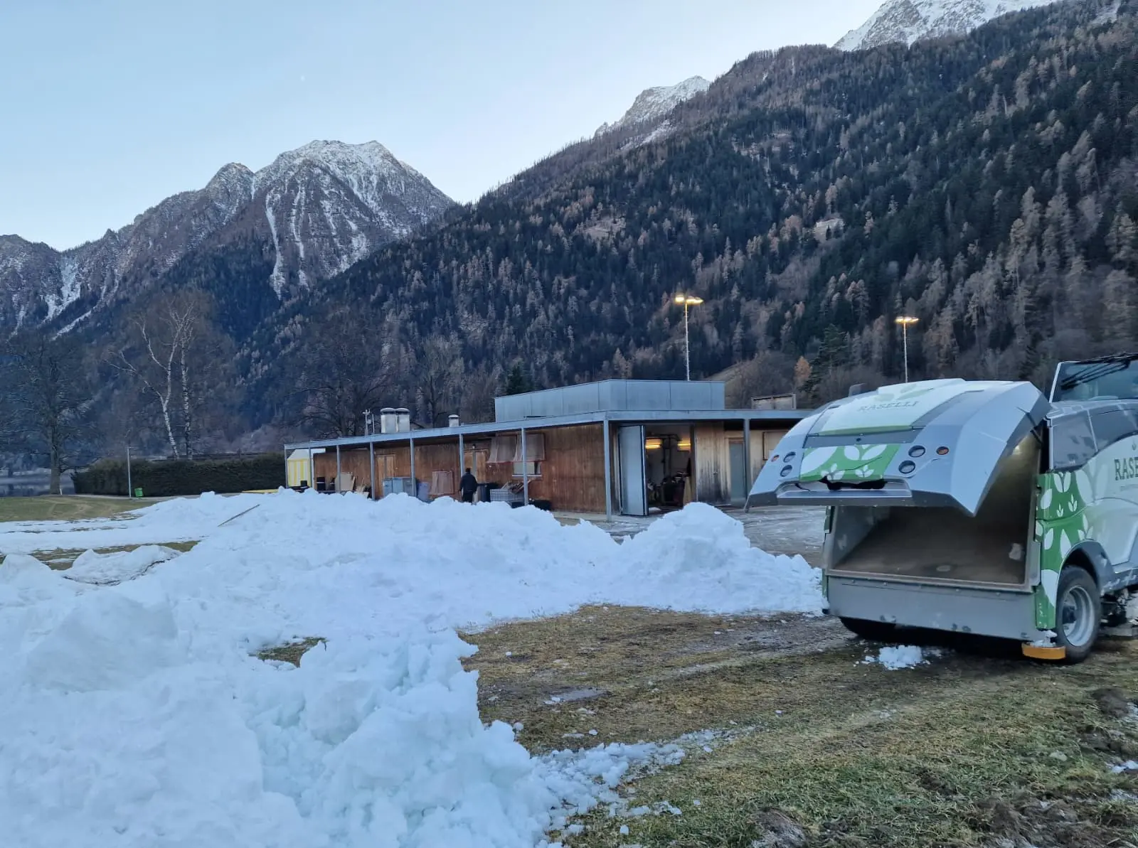 Un'area all'aperto con neve accumulata, un veicolo per la rimozione della neve e montagne sullo sfondo. Un edificio moderno è visibile a sinistra.