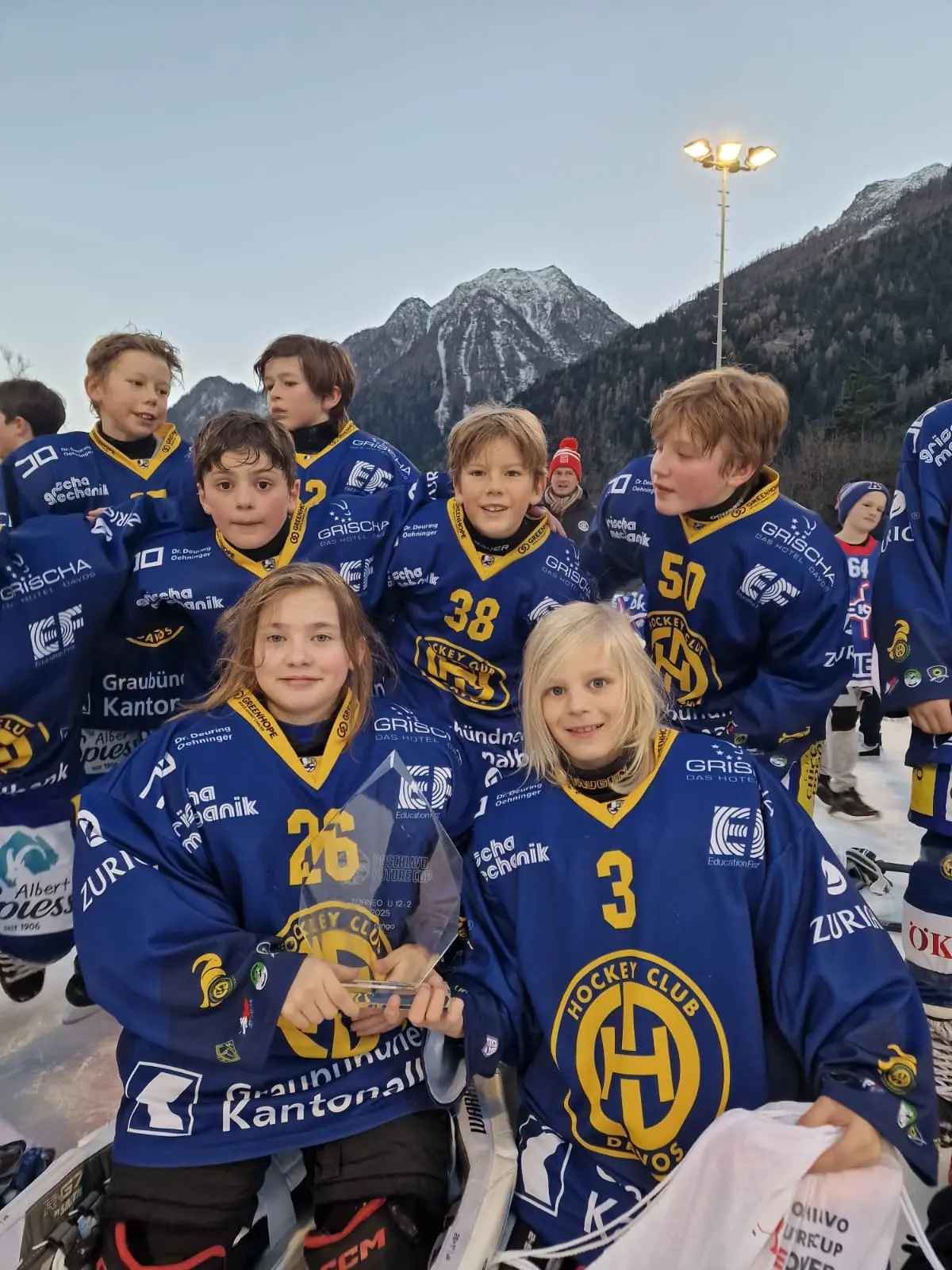 Gruppo di giovani giocatori di hockey su ghiaccio in divise blu del Hockey Club, sorridendo e posando per la foto. Sullo sfondo si intravedono montagne innevate
