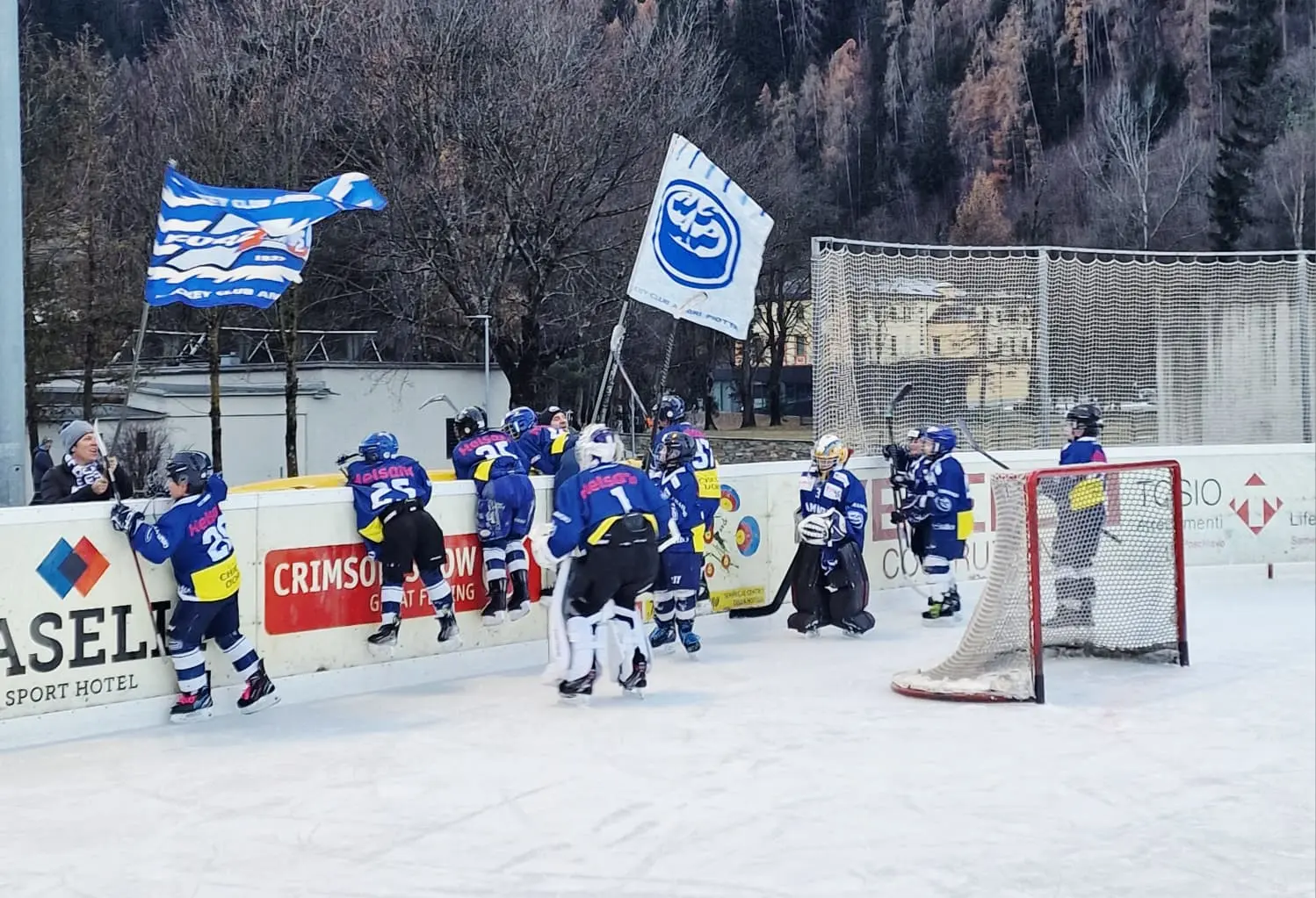 Giovani giocatori di hockey su ghiaccio in uniforme blu stanno entrando e uscendo dal campo, con bandiere che sventolano sullo sfondo. È un pomeriggio di gioco