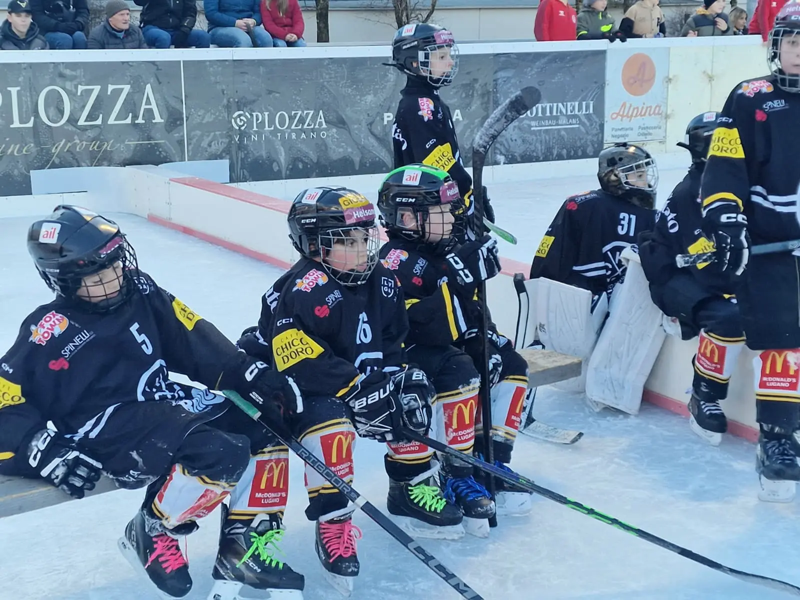 Gruppo di giovani giocatori di hockey su ghiaccio in panchina, vestiti con divise nere. I ragazzi indossano elmetti e guanti, pronti per il gioco