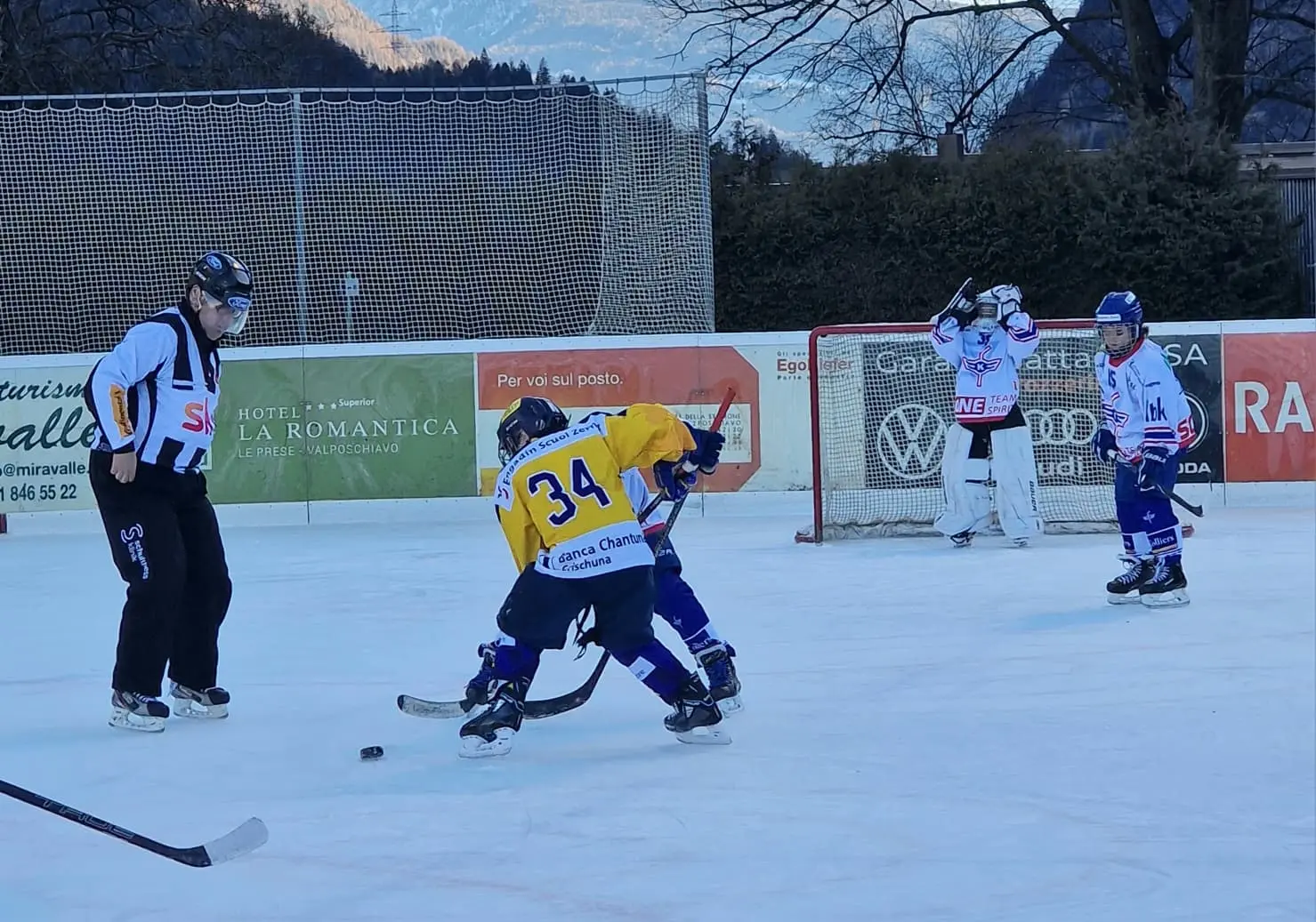 Giovani giocatori di hockey su ghiaccio in un campo all'aperto, mentre si sfidano per il disco. Un giocatore in giallo con il numero 34 è in az